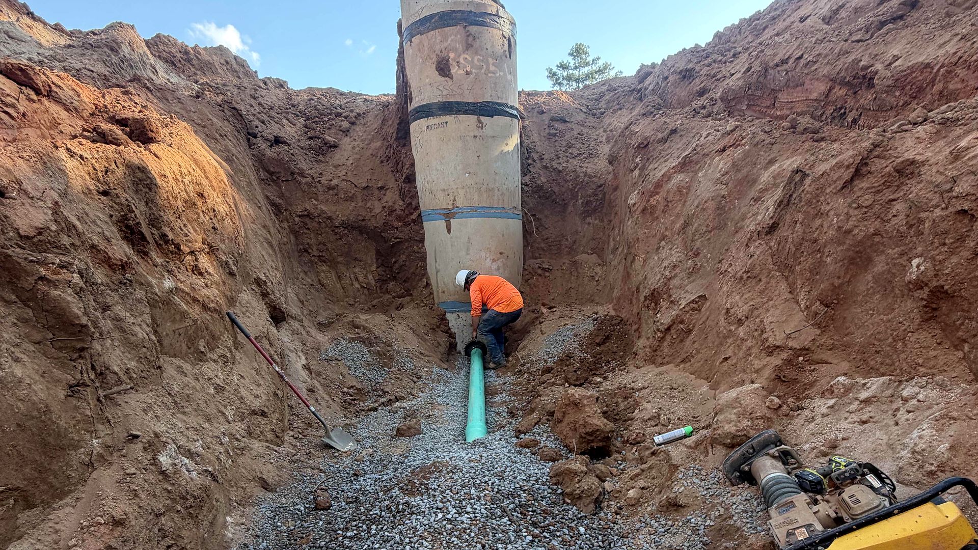 Crew member at the base of a precast manhole with green PVC pipe in a deep trench in Alpharetta, Georgia
